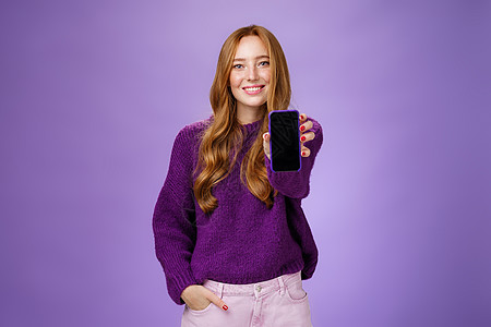 女孩在相机前展示智能手机屏幕 询问朋友的意见 面带乐观和快乐的表情 手牵着口袋 在紫色背景下推销手机或应用程序技术毛衣学生雀斑卷图片