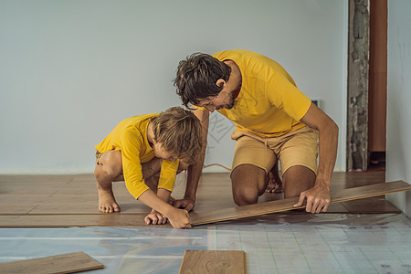 父亲和儿子在温暖的薄膜地板上安装新的强化木地板 复合地板下的红外线地暖系统维修瓦工男人帮助电缆工作服木地板建筑工人房子图片