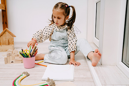 小女孩坐在地板上 用彩色铅笔在草图书中画画图片