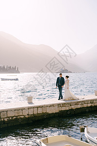 新娘和新郎在黑山佩拉斯特附近岛屿的背景下沿着码头走过港口图片