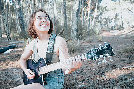 年轻的时髦女人坐在树干上 在公园或花园的背景下弹吉他 少女学习演奏歌曲和创作音乐 爱好 生活方式 放松 仪器 休闲 教育理念热情图片