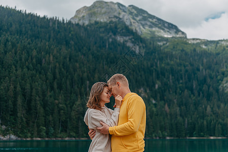 浪漫的新婚夫妇相爱地站在湖边的石岸上 欣赏风景秀丽的山景 新娘和新郎 美丽的白人夫妇在阳光明媚的夏日在湖边拥抱 蜜月的概念蓝色支图片