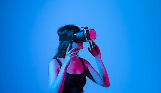 在蓝色背景的霓虹灯下戴着 VR 眼镜玩耍的年轻女子的画像 概念现代小工具和技术 未来的技术概念 虚拟现实游戏视频创新娱乐模拟器头图片