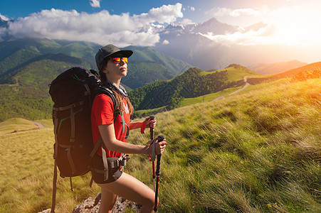 一个背着背包的旅游女孩在山腰欣赏日落 日落时山和云背景中的旅行者 戴墨镜 帽子和背包的女游客图片