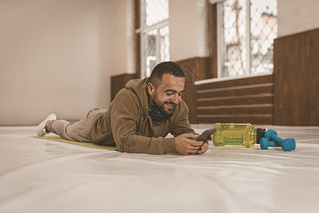 侧面图 穿着运动服的运动型年轻英俊男子躺在地板上的垫子上 观看视频说明或在智能手机上聊天 为隔离者提供的体育视频课程图片