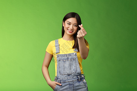 可爱温柔无忧无虑的亚洲女友展示韩国爱情标志 让指心微笑可爱 表现出激情和感情 站在绿色背景的工装裤和黄色 T 恤上 表达同情旅行图片