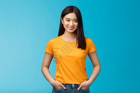 自信 自信 微笑的亚洲女性站在蓝色背景的镜头前 手握牛仔裤口袋 表达自信的氛围 享受看到积极成果的护肤程序 穿黄色 T 恤学生情图片