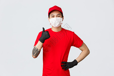 不用担心您的包裹在我们手中是安全的 身着红色制服 防护性医用口罩和手套的自信微笑的亚洲送货员 竖起大拇指表示赞同 说是 保证一切图片