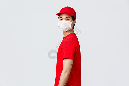 身穿红帽和 T 恤的亚洲送货员的画像 戴着防护性医用口罩 面对镜头 在 covid 19 检疫期间 身着制服的快递员运送包裹 而图片