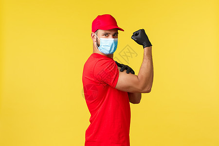 大流行期间的快递 covid19 安全运输 网上购物概念 穿着红色制服和医用面具的强壮的年轻快递员 在黄色背景下展示肌肉 弯曲的图片