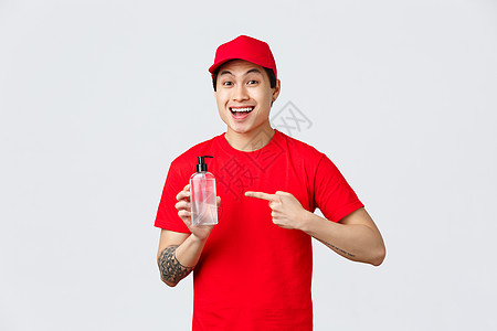 检疫 安全的非接触式交付和购物概念 身穿红帽和 T 恤 面带微笑的亚洲快递员 指着洗手液 提倡安全购物 防止病毒传播的重要性消毒图片