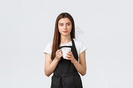 杂货店员工 小型企业和咖啡店的概念 穿着黑色围裙的可爱女咖啡师休息 喝着杯子和微笑的相机 站在白色背景图片