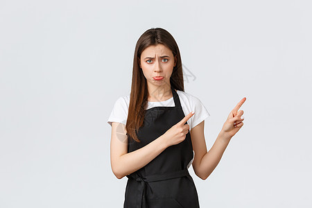 小生意 雇员和咖啡店概念 穿着黑色围裙发怒 皱眉和指向右上角 白背景的年轻女律师协会成员沮丧和失望; 脸红图片