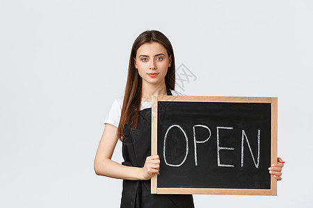 杂货店员工 小型企业和咖啡店的概念 穿着黑色围裙的可爱年轻女店员举着我们开放的标志 告知客人咖啡馆正在工作 白色背景图片