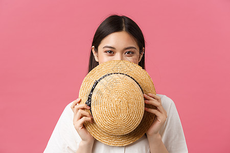 美丽 人们的情感和夏季休闲概念 害羞可爱的日本女孩的特写 在草帽后面遮住脸 微笑着 站着粉红色的浪漫背景 脸红的娇媚图片