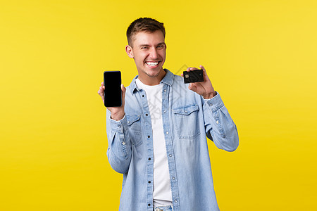 技术 生活方式和广告概念 快乐的金发男子展示信用卡和手机 推荐购物应用程序 并站立在黄色背景上 (请查看InfoFlickr u图片