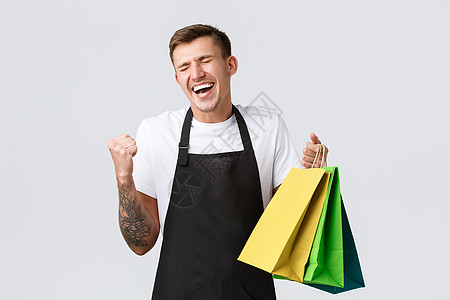 零售店 购物和员工概念 穿着黑色围裙 欢快胜利的推销员 欢欣鼓舞的拳头泵和拿着装有产品的袋子 站在快乐的白色背景中图片