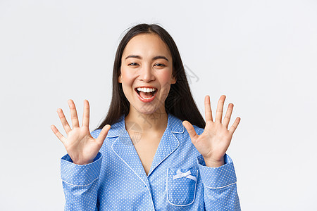 穿着蓝色睡衣的快乐迷人亚洲女性特写镜头 展示十根手指和微笑的白牙 解释主要规则或下订单 站立白色背景 推荐产品图片