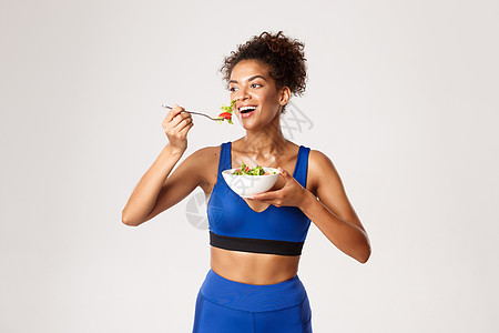 健康生活方式和体育理念 适合身穿蓝运动服装 吃沙拉和微笑 满脸快乐的非洲-美国女运动员 在白色背景中寻找幸福图片