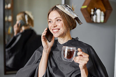 快乐的金发碧眼美女坐在椅子上 在理发厅等美容师时 正在使用智能手机发型头发女孩造型师发型师理发职业客户顾客镜子图片