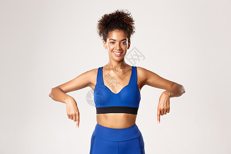 锻炼和健身的概念 身穿蓝色制服的健康年轻女运动员手指向下 微笑着展示标志或横幅 白色背景图片
