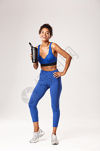 穿着蓝色运动服 饮用水或瓶装蛋白质 在良好的锻炼后笑着微笑 站在白色背景下的全长迷人健康健身女性图片