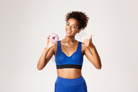 身着蓝色运动服 面带微笑的非洲裔美国女运动员的腰部向上拍摄 竖起大拇指和甜甜圈 微笑着高兴 白色背景图片