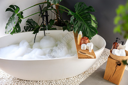现代浴室内部配有白色气泡填充浴缸和龟背竹热带植物 木制家居装饰元素上的生态身体护理产品架子植物群肥皂篮子房间洗发水温泉凝胶洗澡化图片