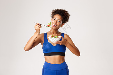 健康的生活方式和运动理念 穿着运动服 白色背景 吃着沙拉 微笑着的美丽非洲裔美国女运动员动机深色运动装皮肤训练福利耐力健身房活力图片