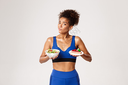 健康生活方式和体育概念 非洲美国女性运动员在饮食 沙拉和蛋糕方面缺乏决定权 试图抵制诱惑 白种背景的白人背景皮肤动机活力衣服福利图片