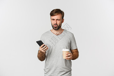 穿着灰色T恤衫 喝着外送咖啡和持有手机 站在白背景上站立的令人失望的皱眉男子照片图片