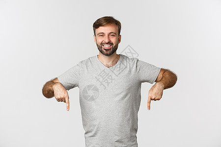 穿着灰色T恤衫的有吸引力的胡须男子肖像 显示徽标 指着手指向下 站在白色背景上学生发型情绪快乐横幅成人微笑男性成功标识图片