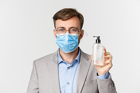 穿着灰色西装和医疗面具的男性老板近距离接近 展示手清洗剂或抗消毒剂 站在白背景上站立在工作 共同生活19和社会疏远的概念中隔离套图片