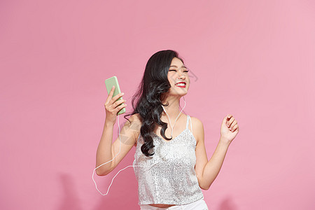 迷人 快乐 情绪化的女人的肖像 戴着耳机 拿着智能手机 在粉红色的墙上摆姿势 从快乐的音乐中微笑图片