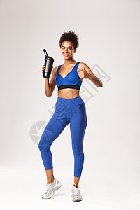 穿着蓝体运动服 饮用水或瓶装蛋白质的全长有吸引力的女性运动员 他们表现了拇指高举 锻炼完后微笑满意 白背景图片