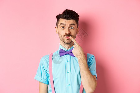 有趣的快乐男人触摸嘴唇 看着摄像头穿孔 选择买什么 站在粉红背景上办公室商业发型吊带裤成人工作室广告情绪微笑成功图片