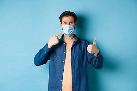 Corona病毒和大流行病概念 年轻健康男子指着医疗面具并举起大拇指 采取预防措施 以从蓝底的Covid19号鱼群中抓获快乐广告图片