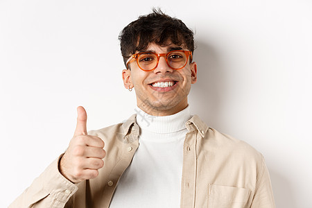 戴眼镜和耳环的帅气现代男人 笑着满足 露出拇指 推荐公司 站在白背景上眼镜赞扬生活发型成人成功情绪学生工作室广告图片