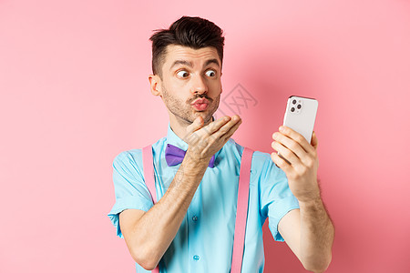 技术概念 有趣的男人在视频聊天时发出空中吻吻 在智能手机相机上吹呼mwah 站在粉红背景上工作室胡须成功办公室广告工具促销吊带裤图片