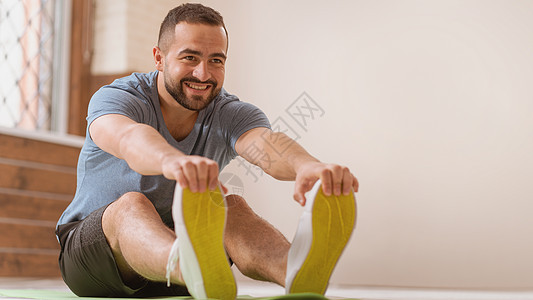 快乐微笑的运动年轻人在家里伸展训练 业余运动员在锻炼活动中坐在垫子上 伸展靠在他的脚趾上 努力工作的人在公寓里锻炼图片
