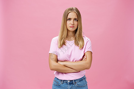 悲伤的嫉妒和失望的阴郁可爱的年轻女人 一头金发 穿着休闲的 T 恤 双臂交叉在胸前 摆出不安的姿势 噘起嘴唇 皱着眉头 在粉红色图片