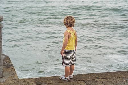 可爱的男孩站在岸边看海浪 看着海浪游客孩子情调岩石童年婴儿天空海滩旅行孤独图片
