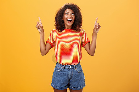 穿着时髦T恤和牛仔短裤的ffro发型 并用有兴趣的眼光仰望在橙色墙上看一看 注   iBAR女性卷曲工作室思维成人手势女孩广告头图片
