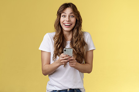 非常高兴热情惊讶快乐可爱的女性女孩在线收到令人愉快的感人消息拿着智能手机广泛微笑看着相机快乐有趣的黄色背景自由职业者情感女朋友工图片