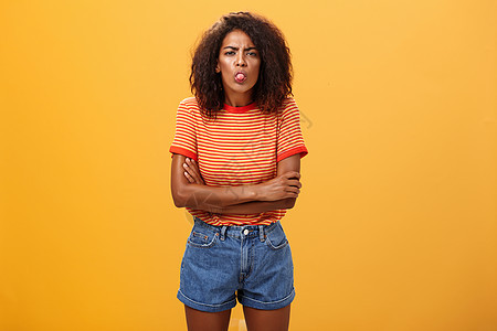 年轻女孩的性格表现不佳 幼稚少女的肖像冒犯或不高兴非洲裔美国妇女 其卷发露出舌头在胸前交叉手臂横跨橙色背景的嘴唇橙子广告发型头发图片