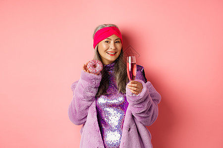 穿着紫花皮大衣 吃甜甜甜圈 喝香槟 在派对上玩得开心 站在粉红色背景之上的优美和时尚的女性促销乐趣成人头巾化妆品食物购物快乐广告图片