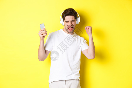 快乐的男子跳舞和耳机听音乐 持有移动电话 站在黄色背景下站立在黄背上音乐商业胡须黑发情绪技术广告手势闲暇成人图片