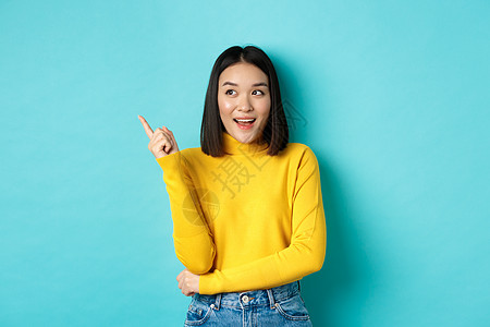 购物概念 穿着黄色毛衣的有吸引力的韩国女孩肖像 展示了在复制空间的升迁提议 指向和看左边满脸笑容 蓝背景商业发型广告黑发促销情感图片