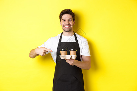 盛着两杯外送咖啡 指着喝酒和微笑的手指头 站在黄色背景的黑色围裙上站立服务手势推销员服务员咖啡店杯子快乐餐厅商业成人背景图片