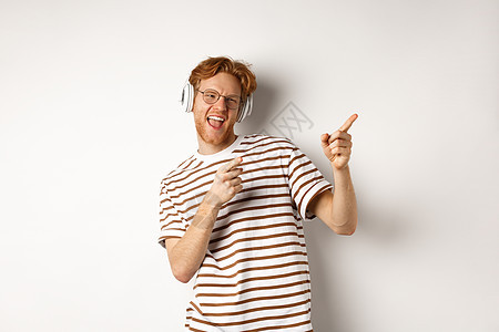 技术概念 快乐的红发男子在耳机中听音乐 跳舞和指右手指 站在白色背景上站立着发型音乐成人男性红色胡须冒充学生工作室头发图片